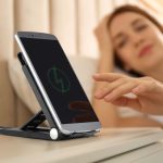A black wireless charger on a bedside table with a smartphone resting on it, featuring a person in the background.