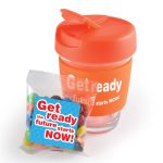 A reusable orange coffee cup with a clear bottom and a pack of colorful M&M's beside it. The cup has a logo.