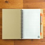 A tan notebook with lined pages and a matching orange pen. The notebook features a spiral binding and a logo.
