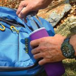 A drink bottle with a purple neoprene sleeve is being placed into a blue backpack among rocks.