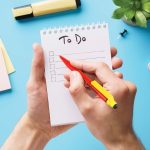 A hand holding a red Chameleon pen writing on a notepad with a "To Do" list, surrounded by stationery items and greenery.