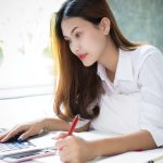 A woman with long, brown hair is writing with a red pen while using a laptop, sitting on a bed.