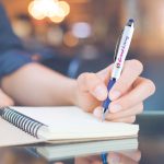 A stylus pen in silver and navy blue held in a hand, writing in a notepad. The pen has a logo on it.