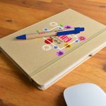 A tan A5 Natura notebook with a floral design and a blue Matador pen on a wooden surface.