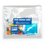 A clear pouch containing colorful candies, a lip balm, a small blue package, and a cleaning cloth, with branding visible.