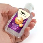 A hand holds a small clear bottle of gel sanitizer with a vibrant purple label featuring bubbles and a logo.
