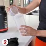 A clear protein shaker with a black lid is being filled with powder. A container of protein powder is nearby.