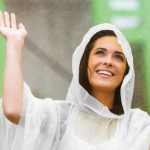 A woman in a transparent white poncho smiles while raising her hand, with rain visible in the background.