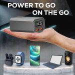 A sleek grey power bank held in a hand, featuring USB ports and an LED display, alongside various tech devices.