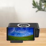 A black wireless charging speaker positioned on a wooden surface, with a smartphone displaying the time.