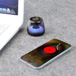 A compact black Bluetooth speaker with RGB LED light next to a smartphone displaying a music app and a laptop.