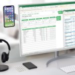 A laptop extension arm in black holds a smartphone above a computer displaying a financial report, with headphones nearby.