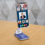 A white foldable phone stand with a blue patterned base, holding a smartphone displaying a video call.