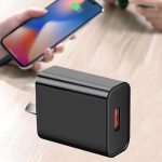A black USB wall charger with a single port, placed on a wooden surface alongside a smartphone.