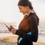 A woman in a black and blue hoodie using a smartphone by the beach during sunset.