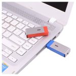 A red and blue Ginan Swivel Flash Drive plugged into a laptop USB port, positioned on a silver laptop keyboard.