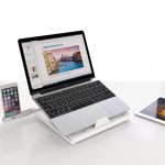 A sleek white foldable laptop stand supports a silver laptop, with a smartphone and tablet nearby.