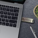 A silver USB flash drive on a grey surface, next to a laptop and a pen, with a plant in the background.