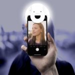 A hand holds a smartphone with a circular white Smiley LED Selfie Light attached, illuminating a person's face in the photo.