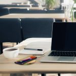 A wireless bamboo fast charger in a natural wood tone on a table near a laptop, notebook, pen, and coffee cup.