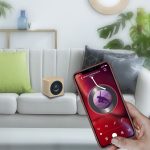A wooden speaker sits on a white couch, with a smartphone displaying a music app in hand, featuring green and beige cushions.
