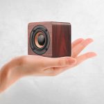 A wooden square speaker in a hand, featuring a circular black speaker grille and a polished finish.