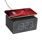 A black alarm clock with a large digital display and a smartphone charging wirelessly on top.
