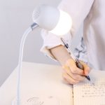 A white wireless charging sound lamp with a curved neck and glowing bulb sits on a desk beside a person writing.