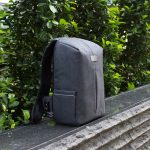 A grey backpack with a sleek design and one side pocket, positioned against a backdrop of greenery.