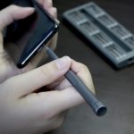 A person holding a grey precision tool near a smartphone, with a tool set displayed in the background.