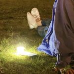 A smartphone torch emits a warm light on the grass, with a person in casual attire sitting nearby.