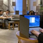 A laptop bag in grey sits beside a person working on a laptop at a café with blurred figures in the background.