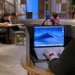 A laptop bag in black with a customer using a MacBook inside a cafe setting, featuring a workspace divider.