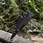 A black multi-tool featuring various tools, including a knife, stably positioned on a wooden surface amidst foliage.