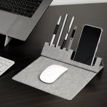 A grey foldable mouse pad with a phone holder and slots for pens, placed beside a laptop and keyboard.