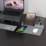 A grey eco mouse pad with a phone stand, holding pens and a smartphone, placed on a desk next to a laptop and keyboard.