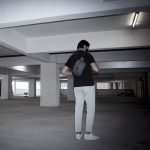 A man in a black shirt and light trousers is carrying a grey anti-theft sling bag in a parking garage.