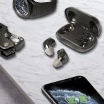 Wireless earbuds in grey resting on a marble surface next to a smartwatch and a smartphone, featuring a logo on the case.