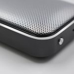 A silver and black Bluetooth speaker with a perforated surface and smooth edges, featuring a small button on the side.