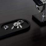 A sleek metallic multi-tool resting on a black tray alongside keys and a ring, set on a polished dark surface.
