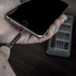 A hand holding a smartphone while using a small black precision screwdriver, with a tool kit in the background.