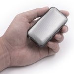 A compact silver wireless speaker held in a hand, featuring a perforated design and a sleek shape.