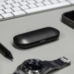 A black charging case for true wireless earbuds rests on a desk next to a watch, a smartphone, and a pen.