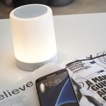 A white touch lamp speaker with a soft glow sits on a table next to a smartphone and a magazine.