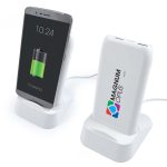 A white wireless power bank and charging station with a smartphone on a stand, displaying a charging battery icon.