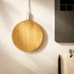 A round bamboo wireless charger in natural wood finish, placed beside a laptop and stationery on a minimalist surface.