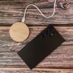 A round bamboo wireless charger in natural wood colour next to a black smartphone on a rustic wooden surface.