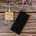 A square bamboo wireless charger in a natural finish beside a black smartphone on a wooden surface.