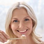 A bamboo toothbrush is held by a woman smiling, with a small amount of toothpaste on the bristles.