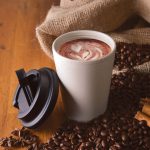 A reusable coffee cup in light beige with a black comfort lid, placed among coffee beans and cinnamon sticks.